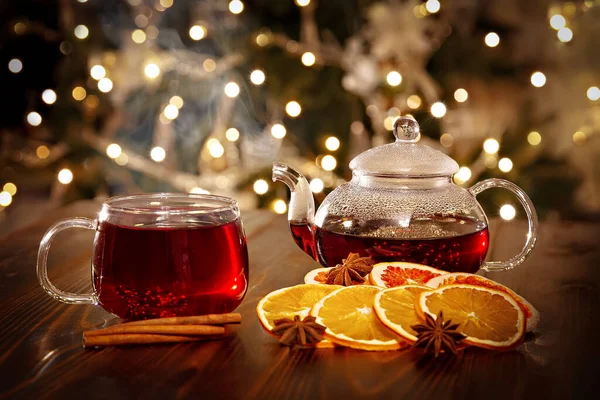 Hot winter drink. Teapot for infusion, transparent cup, cinnamon, dry oranges and star anise on the background of glowing lights