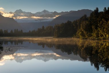 Arkasında dağlar olan bir göl. Su sakin ve gökyüzü berrak. Dağların yansıması suda görülebilir.