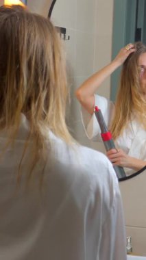 Banyodaki aynaya bakarken uzun sarı saçlarını ütüyle kıvıran bornozlu genç bir kadın. Sabah güzellik rutini, saç bakımı, tımar ve kişisel bakım konsepti..