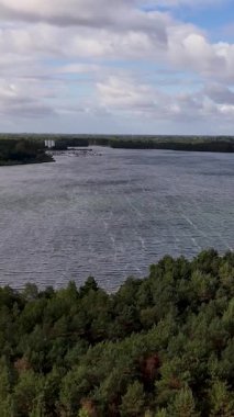 Bulutlu bir gökyüzünün altında yemyeşil bir ormanla çevrili rüzgârlı bir göl manzarası.