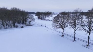 Ağaç çizgisi ve küçük yürüyüşçülerle Karlı Park 'ın geniş bir manzarası var. İnsanlar donmuş arazinin yanında geziniyor.