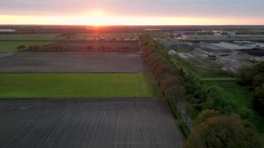 Çiftlik arazisi üzerinde uçan hava görüntüleri ve ufukta güneşin battığı bir sanayi bölgesi.