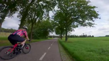 Kırsal bir arazide asfalt bir yolda giden ve beton bir köprünün altından geçen bir sürü bisikletçi.