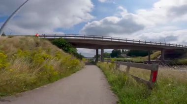 Köprünün altındaki bir kanalın yanındaki kaldırımlı yolda bisiklet sürerken ve ağaçlarla kaplı yeşil bir şeritten geçerken.