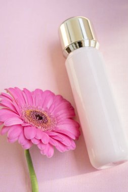 Pembe krem ya da serum kavanozu ya da şeker kutusu ve pembe arka planda altın rengi bir kapak, pembe bir çiçeğin yanında.