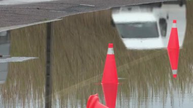 Arabalar kısmen sular altında kalmış bir yolda hareket ederek, yoğun yağmurdan oluşan büyük bir su birikintisine dinamik yansımalar oluştururlar. Dalgalar su yüzeyinde etkileşerek, yağmurlu hava ve kentsel trafiğin atmosferini zorlu koşullarda yakalar..