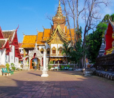 Ağaçlar ve asfalt avlularla çevrili geleneksel bir Budist tapınak binasının dış görünüşü. Mimaride klasik Tayland çatı tasarımları ve dini dekorasyonlar yer alıyor. Otantik gerçek fotoğrafçılık.