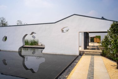 Sakin bir su havuzuna yansıyan eşsiz sanatsal açılımı olan beyaz bir duvarın mimari ayrıntıları. Görüntü yaratıcılığı, zen estetiğini ve modern peyzaj tasarımını aktarıyor. Yüksek kaliteli gerçek fotoğrafçılık..