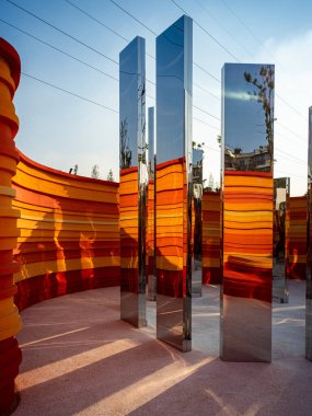 Uzun dikdörtgen aynalı sütunların dikey görüntüsü turuncu eğimli bir duvar manzarasına yerleştirilmiş, gökyüzünü yansıtıyor. Görüntü fütüristik tasarım ve görsel illüzyon taşır. Yüksek kaliteli gerçek fotoğrafçılık..
