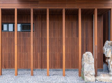 Dikey ahşap kirişleri ve peyzaj taşlarıyla çevrili penceresi olan modern bir binanın ön görüntüsü. Görüntü basitliği ve sürdürülebilir mimari stili aktarır. Yüksek kaliteli gerçek fotoğrafçılık..