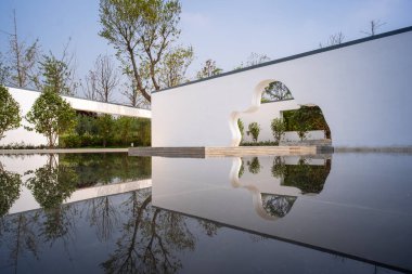 Sanatsal açıkları olan, su havuzuna yansıyan ağaçlarla çevrili beyaz bahçe duvarlarının manzarası. Görüntü, mimari ve doğanın karışımını iletir. Yüksek kaliteli gerçek fotoğrafçılık..