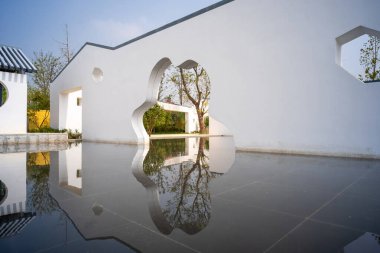 Bulut ve altıgen şekilli açıklıkların bir su özelliğine yansıdığı beyaz mimari bir duvar perspektifi. Görüntü geometrik tasarım ve uzamsal derinlik taşır. Yüksek kaliteli gerçek fotoğrafçılık..