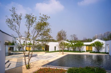 Geniş bir avlu tasarımı, beyaz duvarlar, dikdörtgen bir su havuzu ve mavi gökyüzünün altında manikürlü ağaçlar. Görüntü lüks ve sükunet taşır. Yüksek kaliteli gerçek fotoğrafçılık..