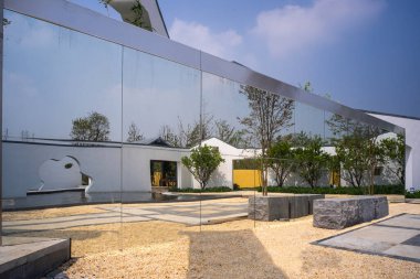 Çevredeki beyaz duvarları ve bahçe ağaçlarını yansıtmak için büyük aynalı bir duvar kullanan modern bir mimari özellik. Görüntü, görsel derinlik ve yenilik taşır. Yüksek kaliteli gerçek fotoğrafçılık..