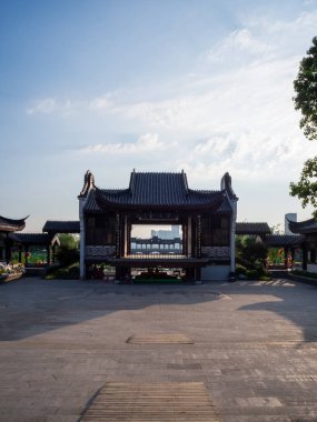 A symmetrical front view of a traditional Chinese gate building or theater stage located in a spacious plaza. The architecture features classic roof tiles and wooden structures under a blue sky.