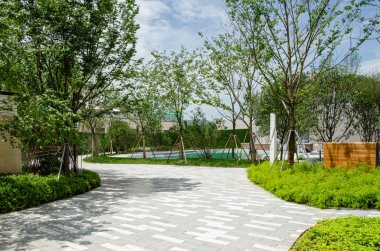 Modern bir konut parkında ya da bahçede dolambaçlı, huzurlu bir kaldırım. Yol yemyeşil ağaçlar ve iyi bakımlı peyzaj ile çevrili, yürümek ve dinlenmek için sessiz bir alan sağlıyor..