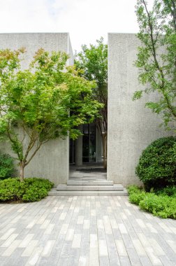 Modern bir minimalist mimari girişin dikey çekimi. Gri beton duvarlar taze yeşil ağaçlar ve kaldırımlı taş kaldırımlarla tamamlanarak temiz ve huzurlu bir şehir tasarımı oluşturulur..
