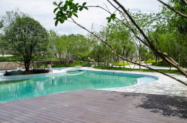 Bereketli yeşil ağaçlar ve ahşap bir güverte ile çevrili temiz bir yüzme havuzu manzarası. Doğal gölgeleme ve berrak su bir bahçe ortamında rahatlatıcı ve ferahlatıcı bir yaz atmosferi yaratır..