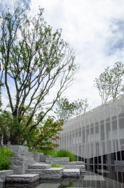 Beyaz yüzgeçli ya da hoparlörlü modern bir binanın dikey görüntüsü. Ön planda, şekillendirilmiş taş bloklar ve ağaçlarla kaplı bir su özelliği mimariye doğal bir dokunuş ekler..