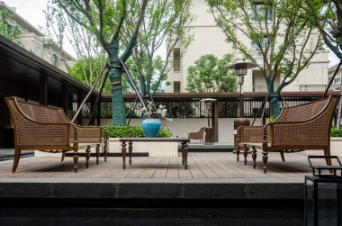 Lüks bir konut bahçesinde sakin bir veranda oturma alanı. Rattan hasır sandalyeler ve bir sehpa ağaçlarla çevrili ahşap bir güvertede, dinlenmek için rahat bir yer yaratıyor..