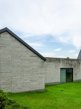 Keskin bir çatı profili ve cam penceresi olan modern beton bir binanın dikey dış görünüşü. Çiğ gri duvarlar mavi gökyüzüne ve yeşil çimlere karşı duruyor..
