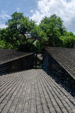 Dikey çekim, gri kiremitli çatıların dokusuna ve geometrisine odaklanıyor. Eğimli açılar ve geleneksel kil maddeler, yeşil ağaçlarla çevrili modern mimari bağlamda kullanılır..