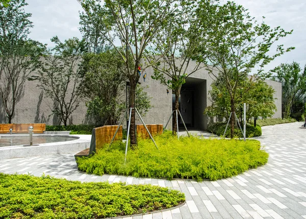 Peyzaj bahçesiyle çevrili modern bir minimalist beton bina girişi. Kavisli kaldırım yolu, taze yeşil zemin örtüsü ve genç ağaçlar temiz ve çağdaş bir mimari ortam yaratıyor..