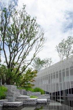 Beyaz yüzgeçli ya da hoparlörlü modern bir binanın dikey görüntüsü. Ön planda, şekillendirilmiş taş bloklar ve ağaçlarla kaplı bir su özelliği mimariye doğal bir dokunuş ekler..