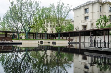 Büyük bir yansıtıcı su havuzu olan modern bir konut avlusunun simetrik görüntüsü. Ağaçlar su özelliklerinin içine dikilir ve etrafındaki binalar sakin karanlık yüzeye mükemmel bir şekilde yansıtılır..