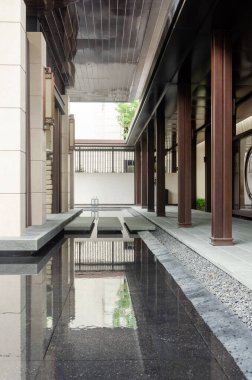 Sakin bir su özelliğinin yanında uzanan modern bir mimari koridorun yakın çekim görüntüsü. Tasarım, geleneksel Asya elementlerini çağdaş malzemelerle harmanlayarak sakin bir yerleşim ortamı yaratıyor..