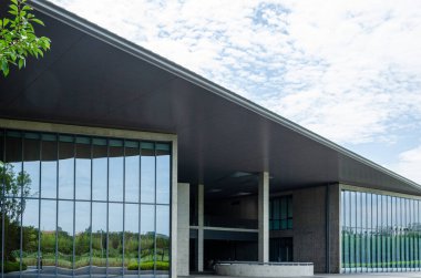 Geniş, düz bir çatı ve cam cepheli modern bir binanın alçak açılı görüntüsü. Mimari çizgiler bulutlu bir gökyüzüne karşı güçlü bir geometrik kompozisyon oluşturuyor..