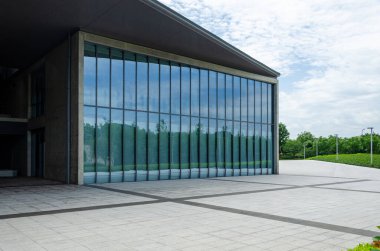 Cam bir kutuya benzeyen, cam perdeli, modern bir bina. Uzak ağaçlarla kaplı asfalt bir plazada yer alan bina, bulutlu gökyüzünü ve çevreyi yansıtıyor..