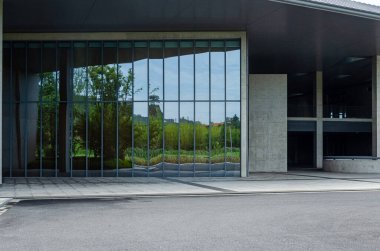 Geniş cam duvarları ve koyu beton çatılı bir çıkıntısı olan modern bir kamu binasının girişi. Tasarım minimalist ve işlevsel, pencerelerdeki yeşilliği yansıtıyor..