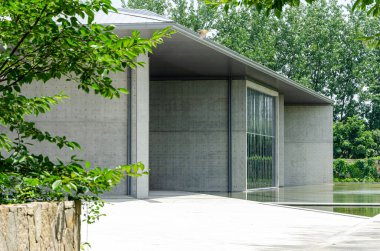 Modern beton bir binanın dış görünüşü, muhtemelen yeşil ağaçların arasına yerleştirilmiş bir kütüphane ya da galeri. Tasarımda ham beton duvarlar, büyük bir cam pencere ve düz bir çatı çıkıntısı yer alıyor..