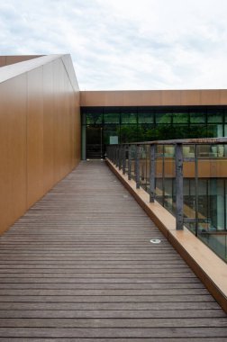 Modern bir binanın yan tarafına uzanan tahta bir tahta kaldırım ya da güverte yolu. Mimaride sıcak renkli kaplama panelleri, cam korkuluklar ve temiz çizgiler yer alıyor ve çağdaş bir tasarım yaratıyor..