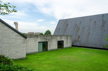 Ham beton duvarlar ve büyük eğimli gri kiremit çatılı modern bir mimari binanın dış görünüşü. Manikürlü yeşil bir çimen, endüstriyel malzemelerle çelişen bir avluda duruyor..