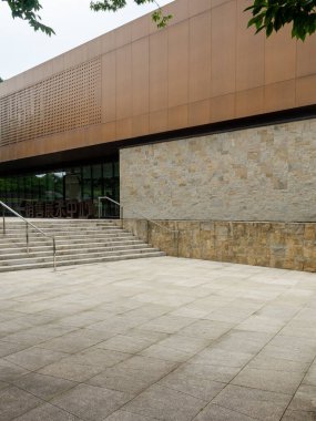 Sıcak ahşap paneller, cam pencereler ve desenli taş bir duvarın kombinasyonundan oluşan modern bir binanın dışı. Taş basamaklar girişe çıkıyor, halka açık bir yer yaratıyor..