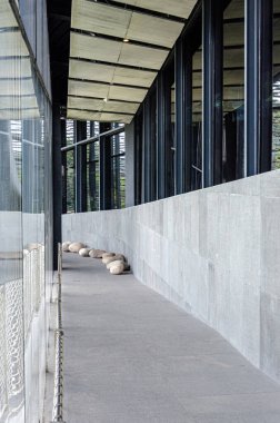 Kıvrımlı cam bir duvar ve gri taş kaplamalı gösterişli modern bir koridor. Taş heykeller ya da koltuklar zemin boyunca yerleştiriliyor ve doğal ışıkla dolu minimalist ve sanatsal bir yol oluşturuyor..