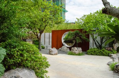 HANGZHOU, ÇİN - 3 Temmuz 2017: Modern Çin tarzı yerleşim bölgesinde peyzaj yapılmış bir bahçe. Daire şeklinde ay kapısı, şekilli bonsai ağacı ve beyaz çakıl zemini olan paslı Corten çelik bir duvara sahiptir..