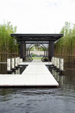 HANGZHOU, ÇİN - 3 Temmuz 2017: Su birikintisinden geçen beyaz taş yol modern bir siyah metal pavyona çıkıyor. Yapıda geleneksel dairesel bir ay kapısı bulunur ve bir bambu korusu tarafından desteklenir..