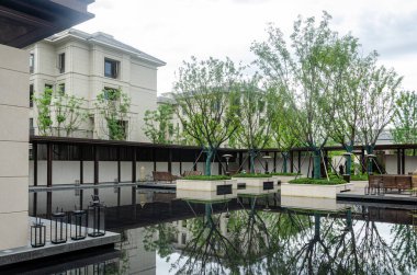 HANGZHOU, ÇİN - 04 Temmuz 2017: Koridor boyunca sakin su, yeşil ağaçlar ve açık hava oturma alanlarının yer aldığı modern bir yerleşim yeri bahçesi manzarası. Apartman binalarının bej rengi taş ön cephesi arka planda görünüyor..