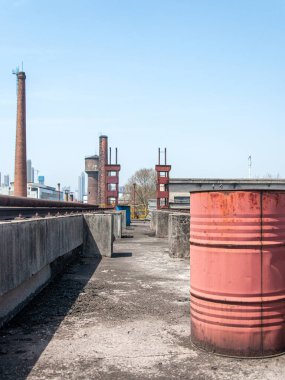 Arka planında beton yapılar ve bacalar olan paslı bir kırmızı metal yağ varili ya da varili olan terk edilmiş bir sanayi çatısı manzarası..