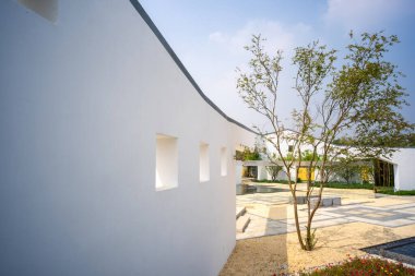 Çağdaş manzara mimarisi, kare pencereli beyaz kıvrımlı bir duvar ve mavi gökyüzünün altındaki çakıllı bir avluda bir ağaç içerir..