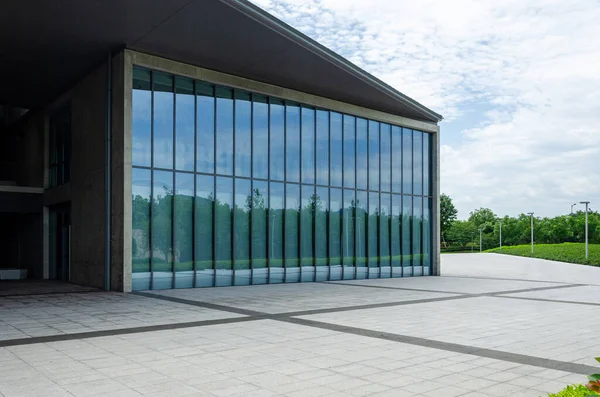 Cam bir kutuya benzeyen, cam perdeli, modern bir bina. Uzak ağaçlarla kaplı asfalt bir plazada yer alan bina, bulutlu gökyüzünü ve çevreyi yansıtıyor..