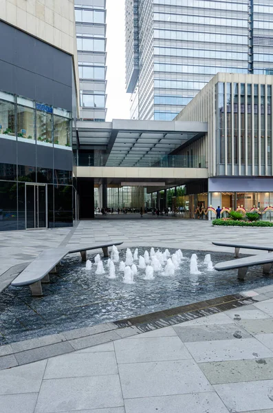 Modern bir ticari yaya caddesinde ya da alışveriş merkezinde küçük bir çeşmenin dikey görüntüsü. Bölge taşla kaplanmış ve cam binalarla çevrili. Temiz bir şehir ortamı yaratıyor..