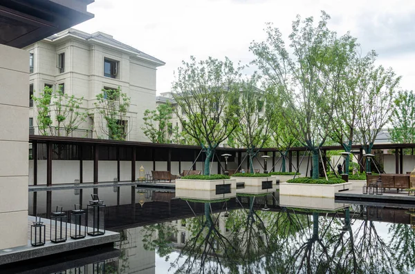 HANGZHOU, ÇİN - 04 Temmuz 2017: Koridor boyunca sakin su, yeşil ağaçlar ve açık hava oturma alanlarının yer aldığı modern bir yerleşim yeri bahçesi manzarası. Apartman binalarının bej rengi taş ön cephesi arka planda görünüyor..