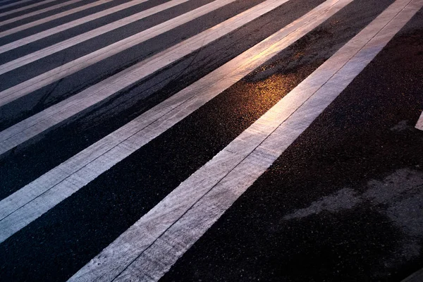Siyah asfalt bir yolda beyaz yaya zebranın yüksek açılı görüntüsü altın güneş ışığıyla aydınlatılmış desen oluşturur..
