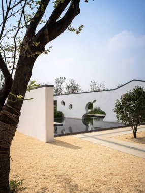 Beyaz duvarları, düzensiz pencereleri, ağaçları ve mavi gökyüzünün altındaki su birikintisi olan modern Çin tarzı bir bahçenin huzurlu manzarası..