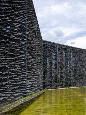 Yeşil bir su havuzu üzerindeki kablolara asılmış binlerce gri çatı fayansının oluşturduğu devasa bir duvarın soyut mimari görüntüsü.