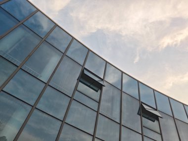Bulutlu gökyüzünü yansıtan kavisli mavi cam perde duvarlı modern bir şirket binasının mimari ayrıntıları.